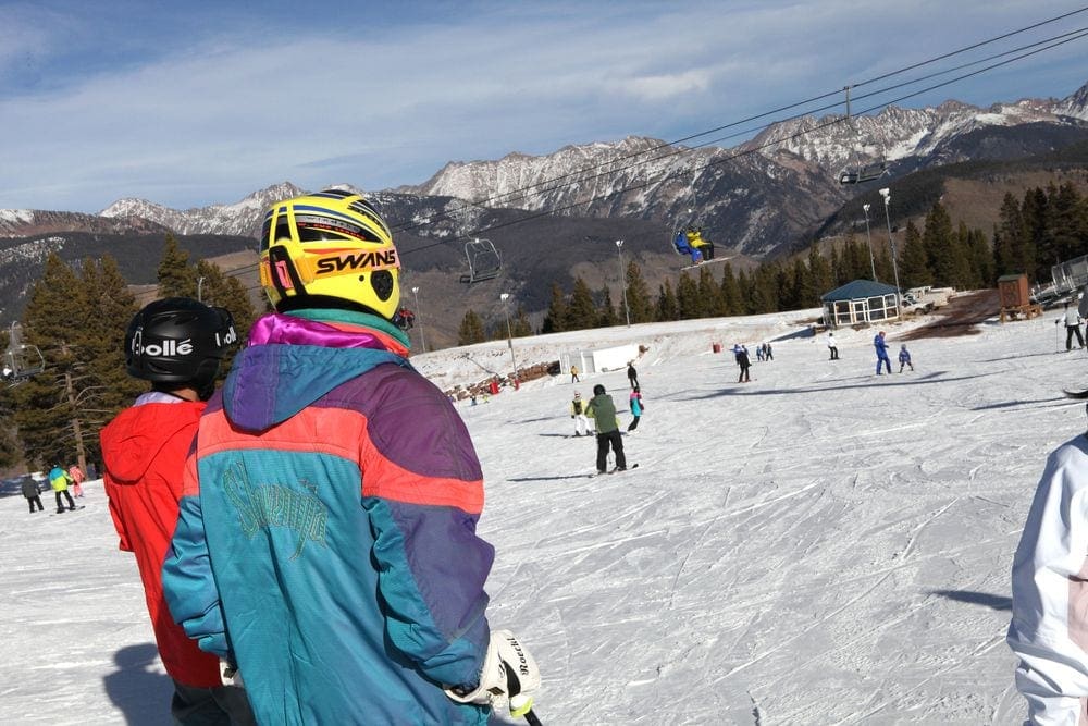 Two skiers in snow-gear and helmets look on at the slopes at Adventure Ridge, a fun place to visit in Vail in the winter with kids, with a ski lift and other skiers in the background.