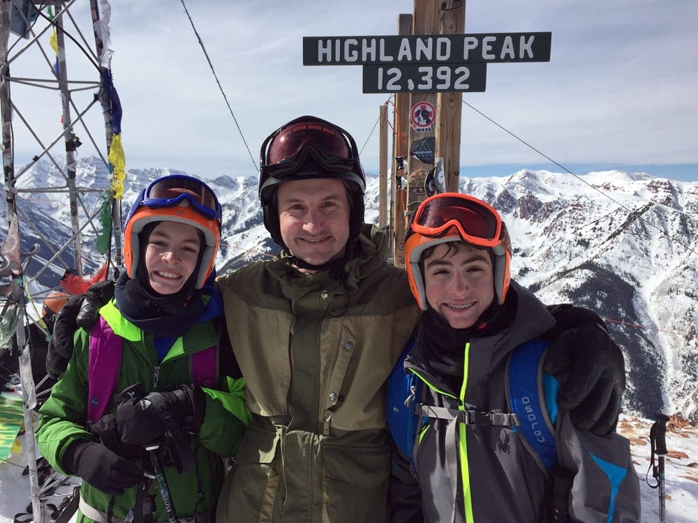 A dad and his two kids in snow gear stand in front of a sign reading "Highland Peak, 12,392" at Aspen/Snowmass, one of the best Colorado ski resorts for families.
