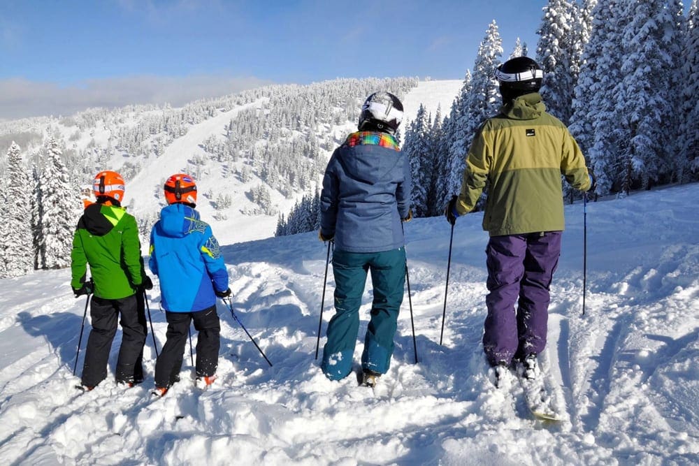 A family of four, all in skis and ski gear, looks out at the runs at Vail on a sunny day.