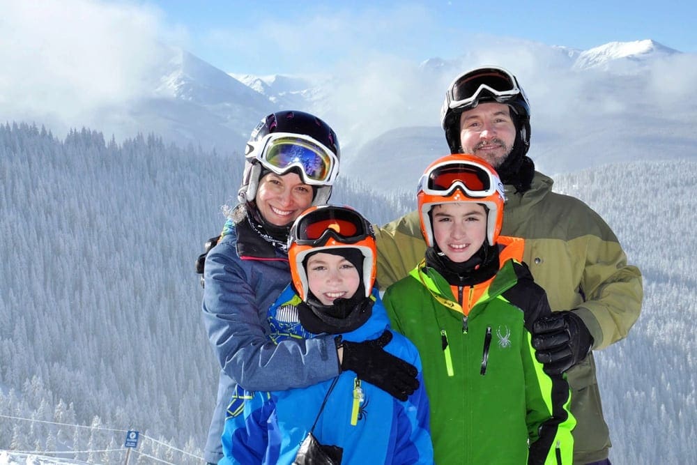 A family of four, all wearing snow gear, smiles while standing on the top of a slope at Vail, one of the best Colorado ski resorts for families.