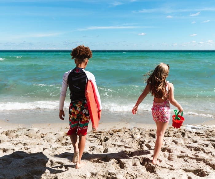 A young black boy carrying a body board and a young white girl carrying a pail walk toward the ocean in Florida near the Photo Courtesy: The Acqualina Resort.