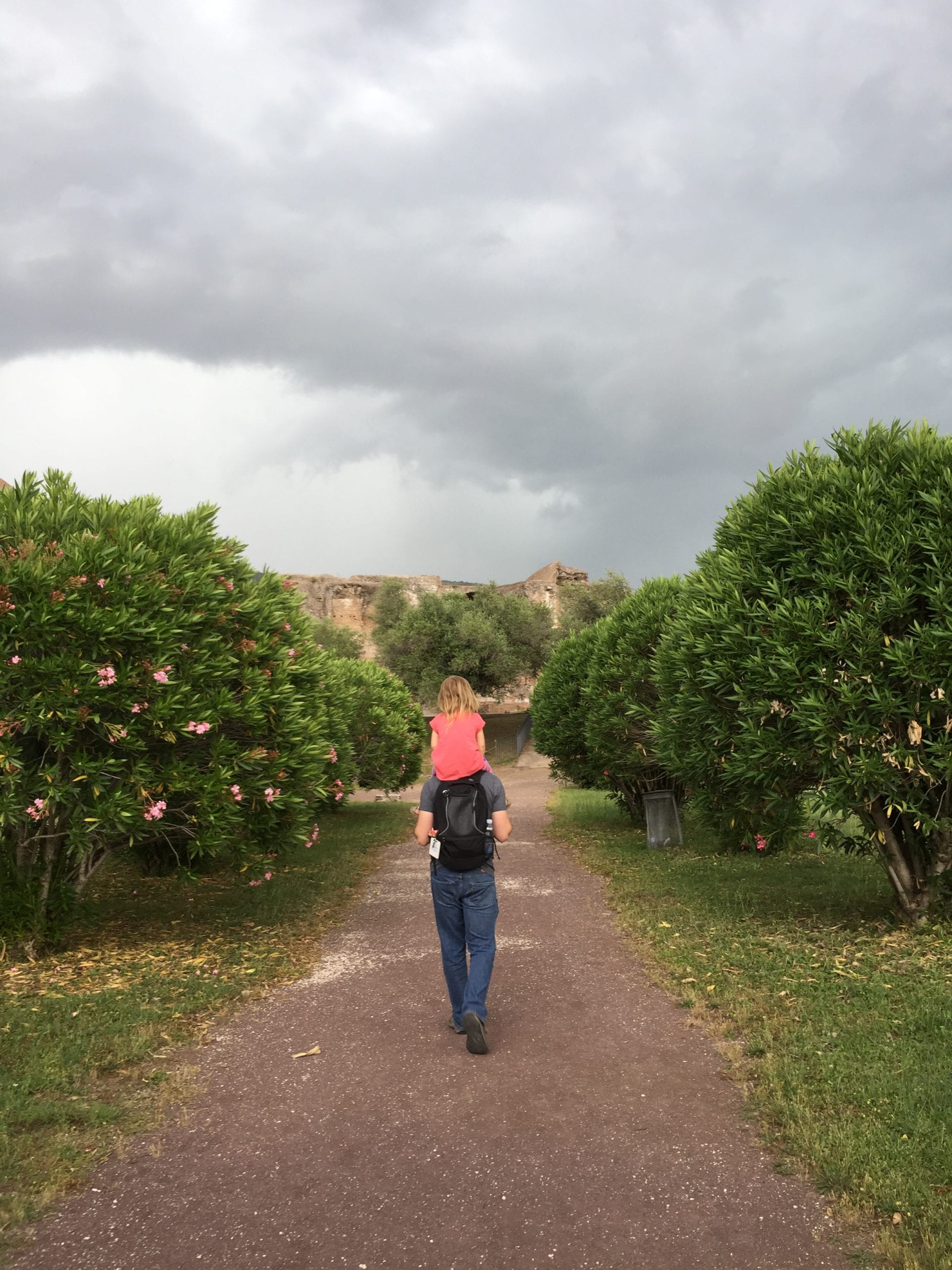 A young girl rides on her father's shoulder inside Hadrian's Villa near Rome, walking amongst the fruit trees.