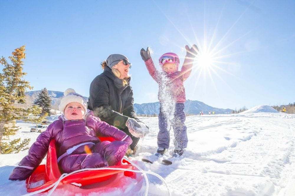 A young baby sits on a red sled with mom and older sister nearby.