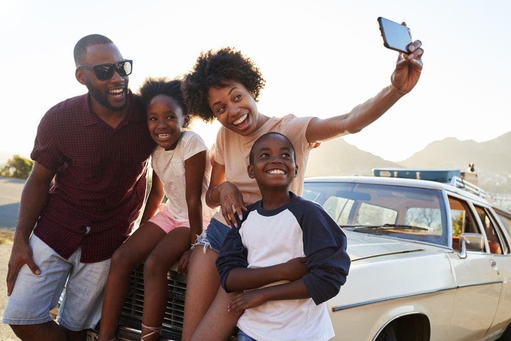A black mom holds out her hand with an iPhone to take a selfie of her family, emphasizing the journey is one of the best tips for taking a family vacation on a budget.