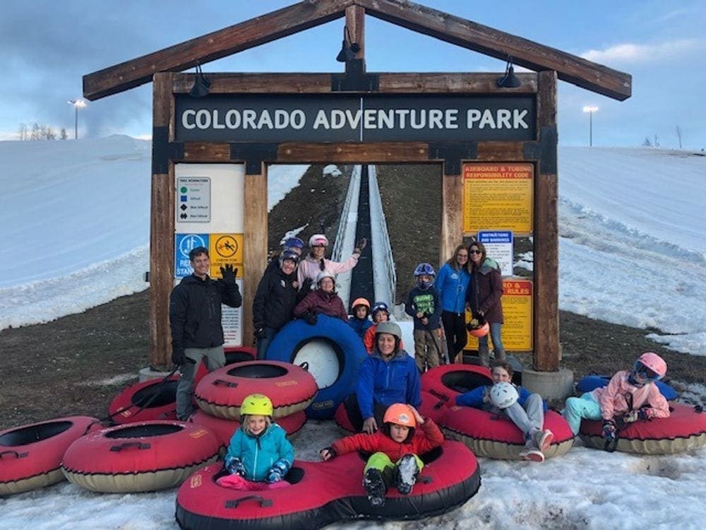 A number of adults and kids on snow tubes stand in front of a sign reading "Colorado Adventure Park", one of the best Colorado snow tubing spots!