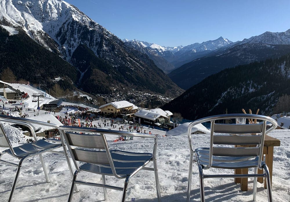 Three empty chairs sit in the snow looking toward a busy ski chalet in Courmayeur.