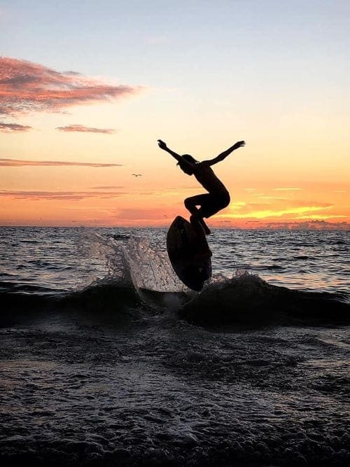 A woman on a surfboard leaps from the water at sunset.