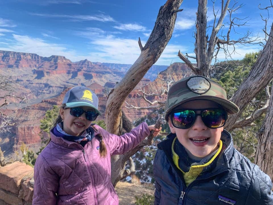Two kids wearing winter gear, hats, and sunglasses smile with an expansive view of Grand Canyon National Park behind them. This is one of the best spring break family destinations.