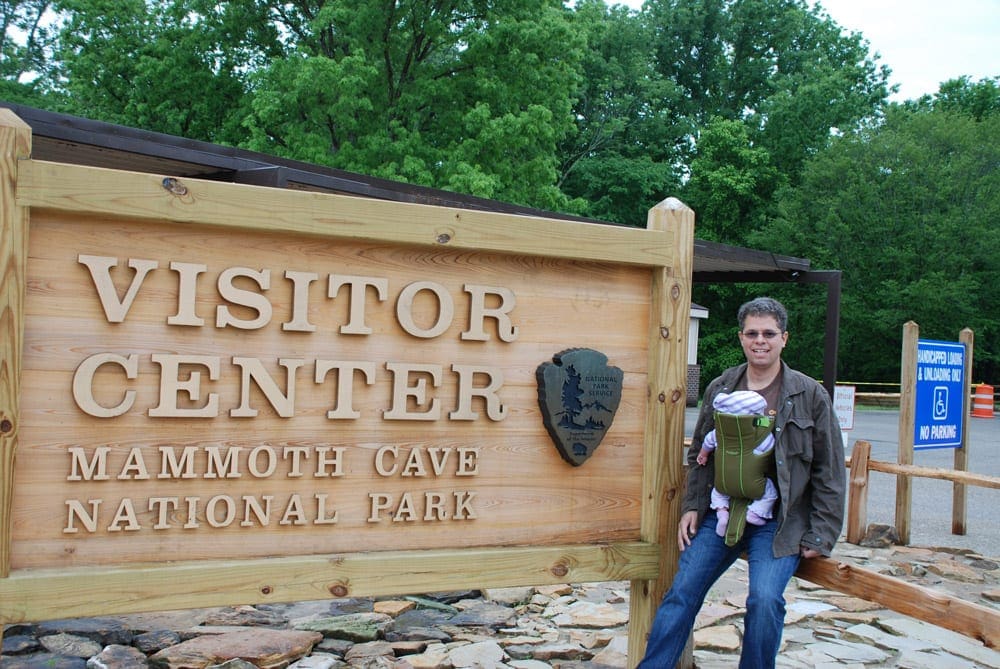 A dad wearing his young infant on his chest stand next to the Visitor Center sign for Mammoth Cave National Park during the winter.