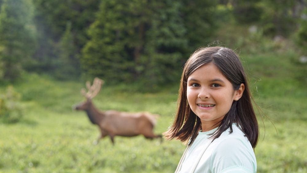 A young girl smiles at the camera while an elk runs in the background.