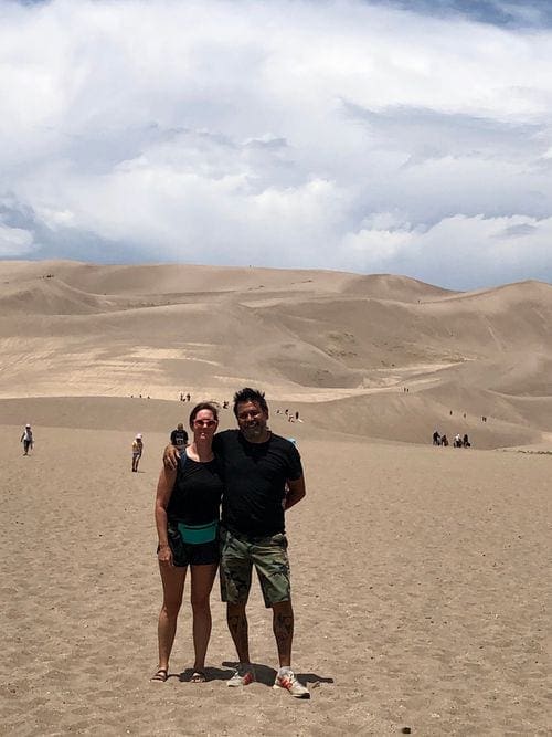Two adults stand together in sand dunes.