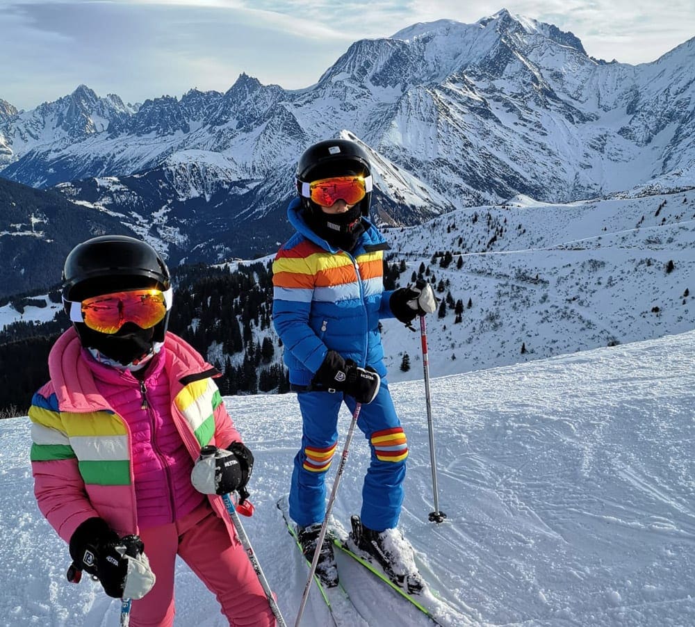 Two kids in full colorful ski gear, including ski goggles and helmets. Proper ski clothing is a key component of our guide to ski gear for kids.
