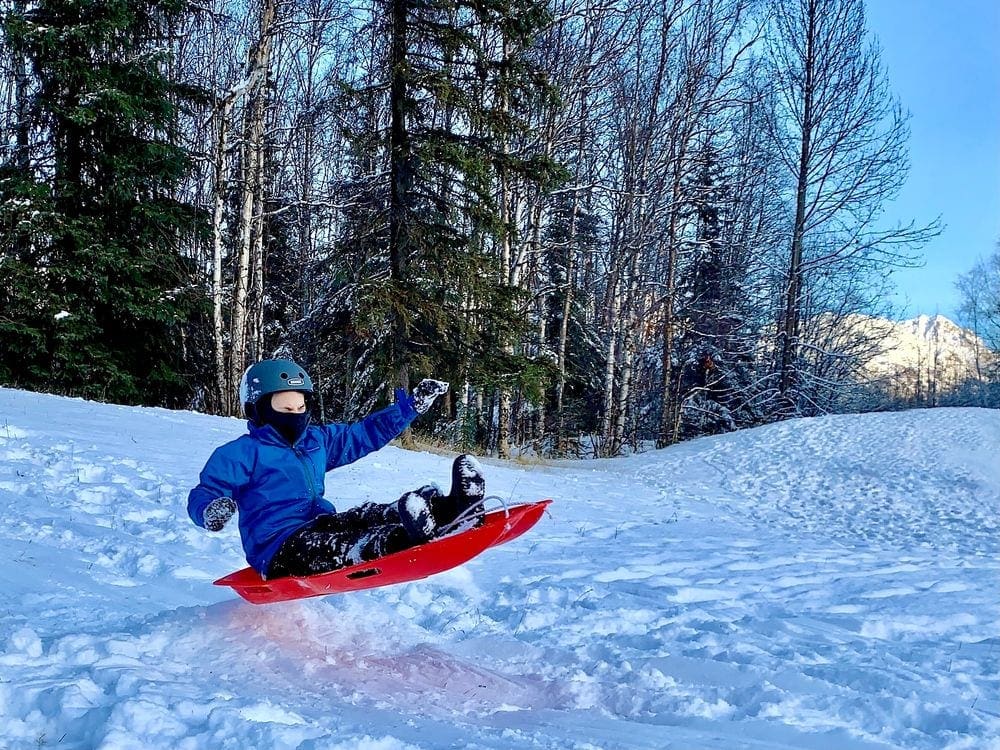 A young boy wearing a blue helmet and a blue coat sits on a red sled airborne, as it goes over a small snow mound.