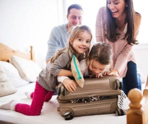 A family of four sits on a bed packing a suitcase.