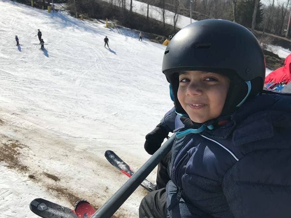 A young boy smiles while riding a chair lift at Mountain Creek, one of the best ski resorts near NYC for families, skiers are seen down below.