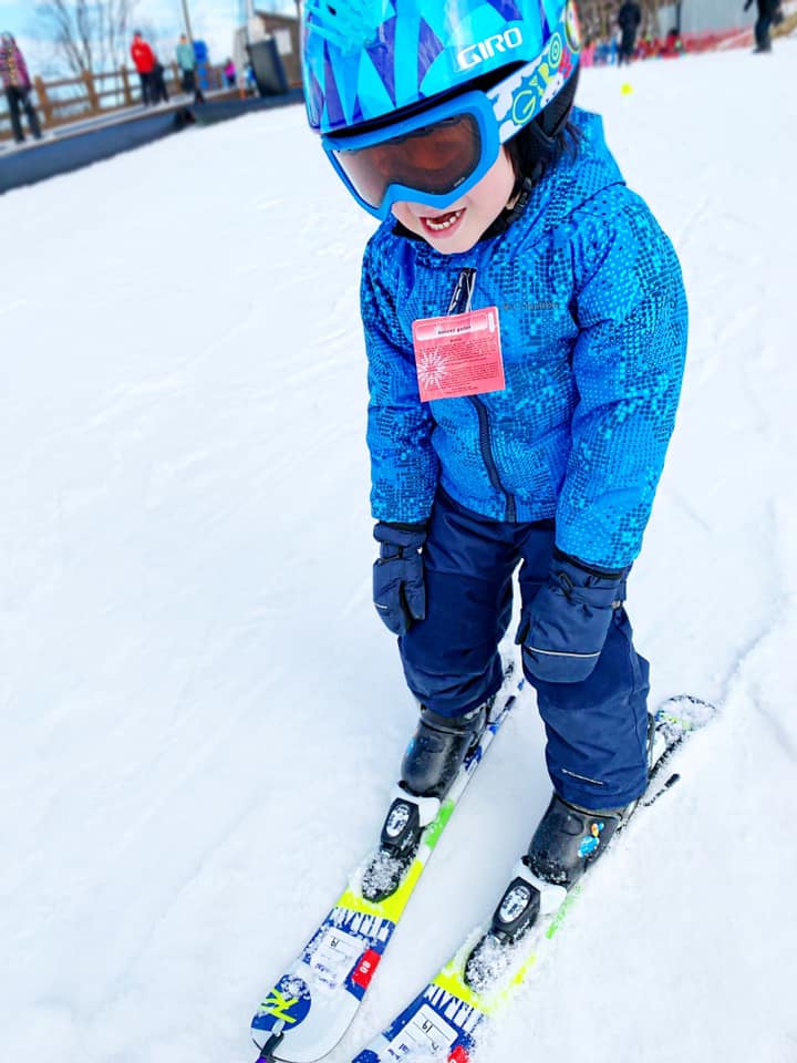 A young boy, wearing a full blue snowsuit and helmet, happily skis at Mount Peter.