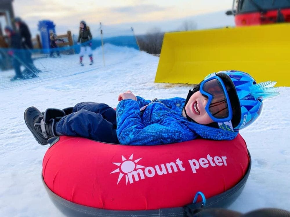 A young boy wearing ski goggles smiles broadly while laying on a red tube at Mount Peter. Proper ski goggles are a key component of our guide to ski gear for kids.