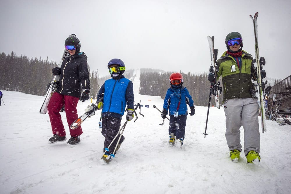 A family of four outfitted in snow gear, all holding their own skis and poles, walks along at Monarch Mountain, best small ski resorts in Colorado for families.