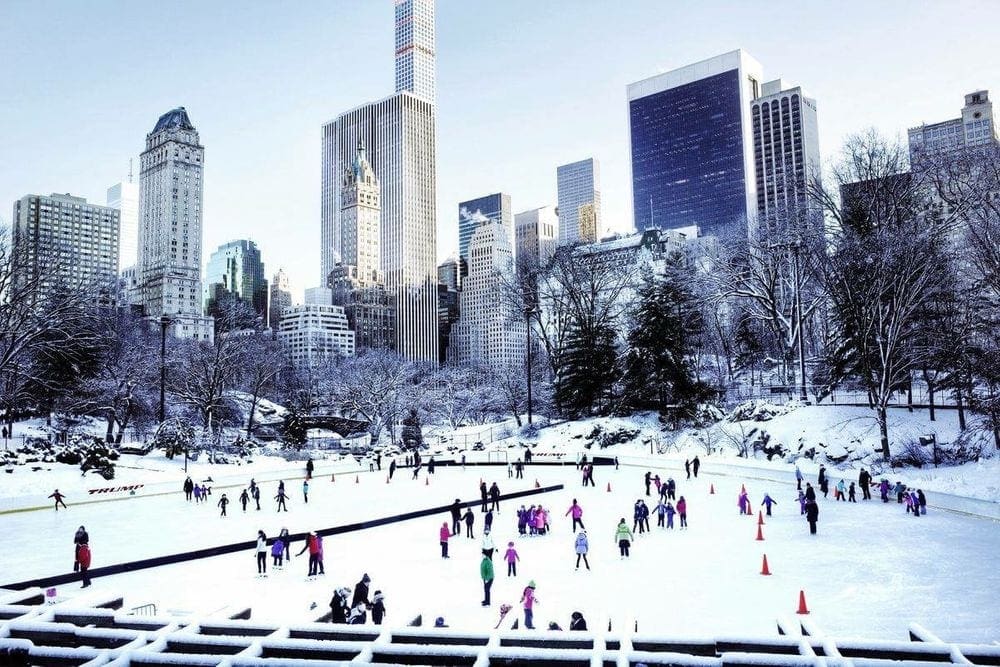 A view of the Wollman Rink on a snow-covered day featuring a number of skaters, and the NYC skyline behind them.
