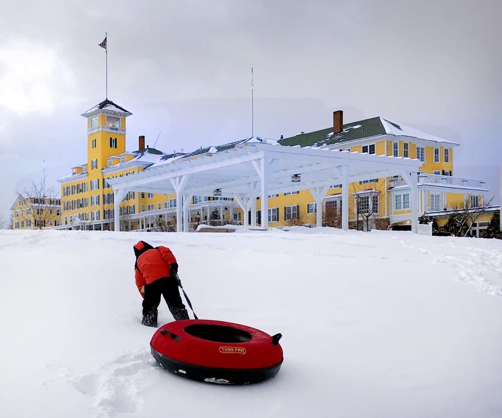 A young boy wearing a red coat pulls a red snow tube across the snow in front of a winter resort.