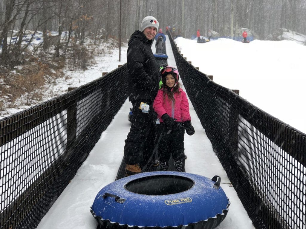 A young girl stands with her dad, holding a snow tube, while riding the conveyer belt at Rocking Horse Ranch, one of the best family resorts near NYC for a winter getaway.