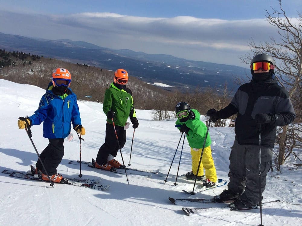 A parent and three kids stand at the top of a ski run at Bromley.