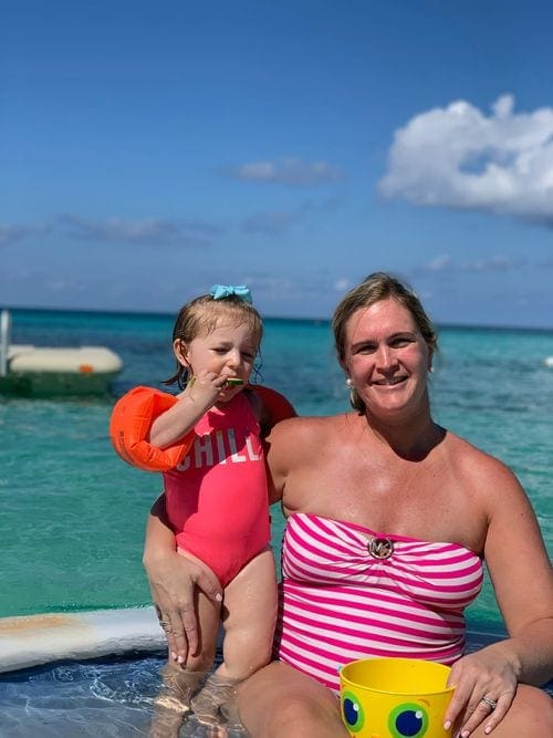 A mom wearing a pink and white swimsuit puts her arm around her toddler daughter while swimming at the Grand Cayman Marriott Beach Resort.