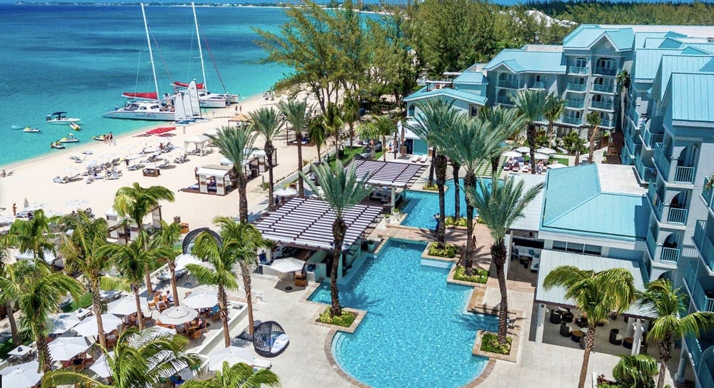 An aerial view of The Westin Grand Cayman Seven Mile Beach Resort & Spa, featuring a pool, palm trees, sail boats, and large hotel buildings.