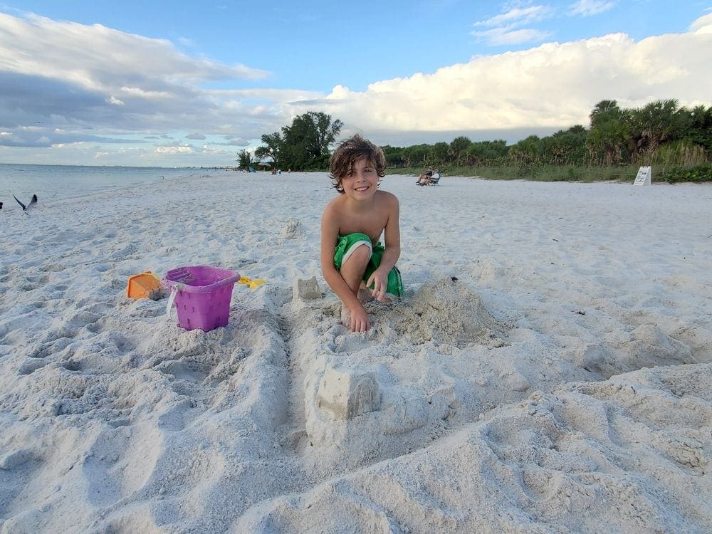 A young boy plays in the sand on a beach near Naples, along side him is a puple bucket and yellow shovel for building sand castles.