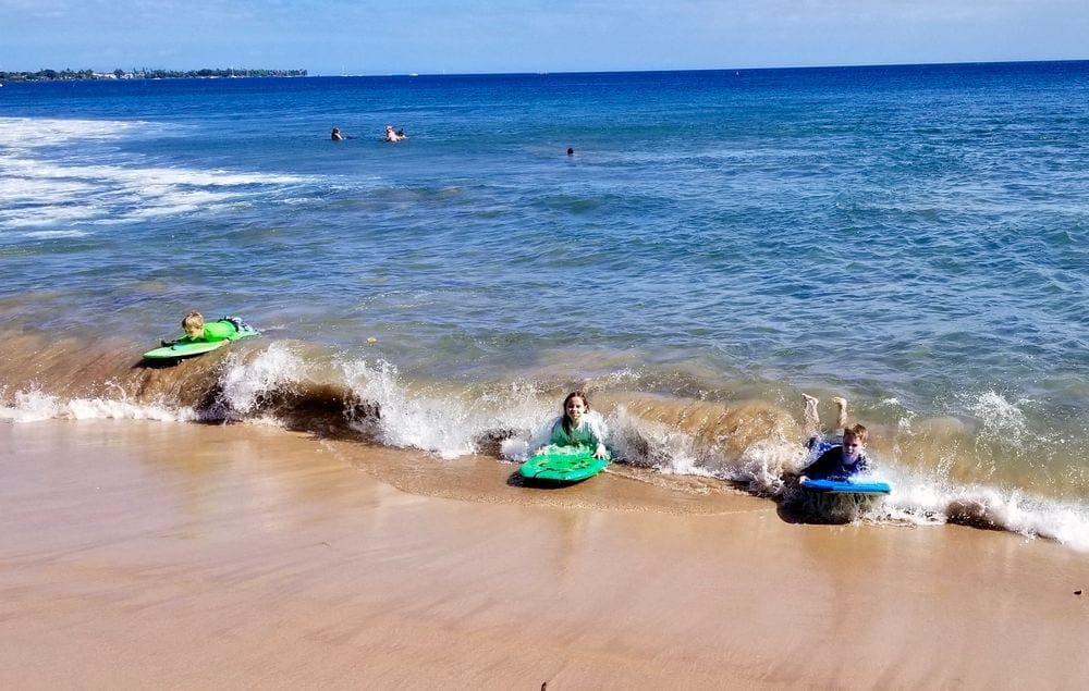 Three kids ride on paddleboards along a small wave on the beach in Maui.