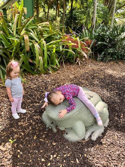 A young girl hugs a large frog statue while her sister looks on at the Miami Zoo.