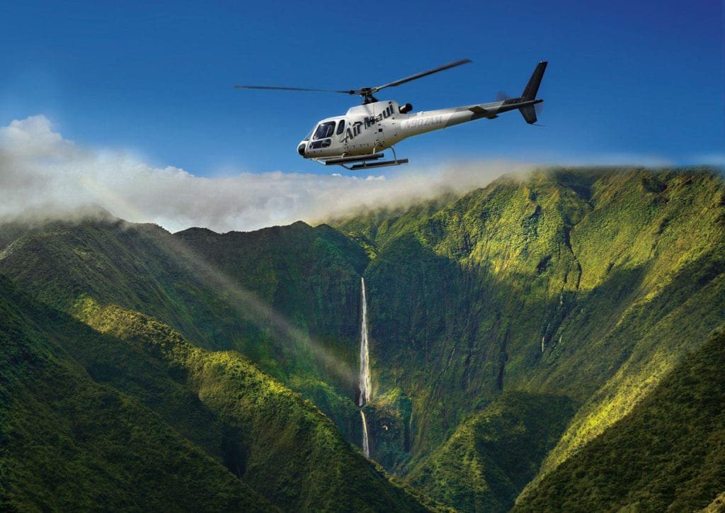 A white helecopter flies over lush mountains in Maui.