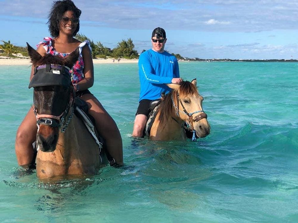 A black woman and a white man ride horses in the water off the coast of Turks & Caicos.