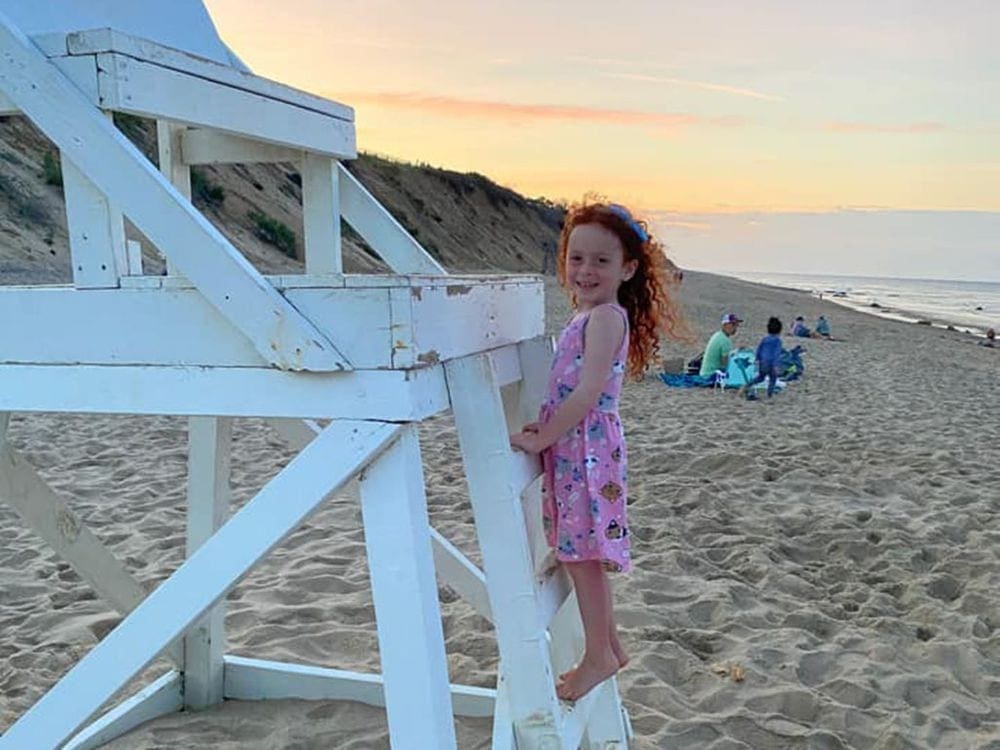 A young girl with red hair and a pink dress climbs a life guard chair on Coast Gaurd Beach in Cape Cod, one of our recommended kid-friendly U.S. beaches for families.