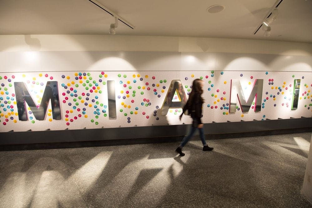 A blurred woman walks across a sign within the Phillip and Patricia Frost Museum of Science, reading "MIAMI" along a wall of the museum.