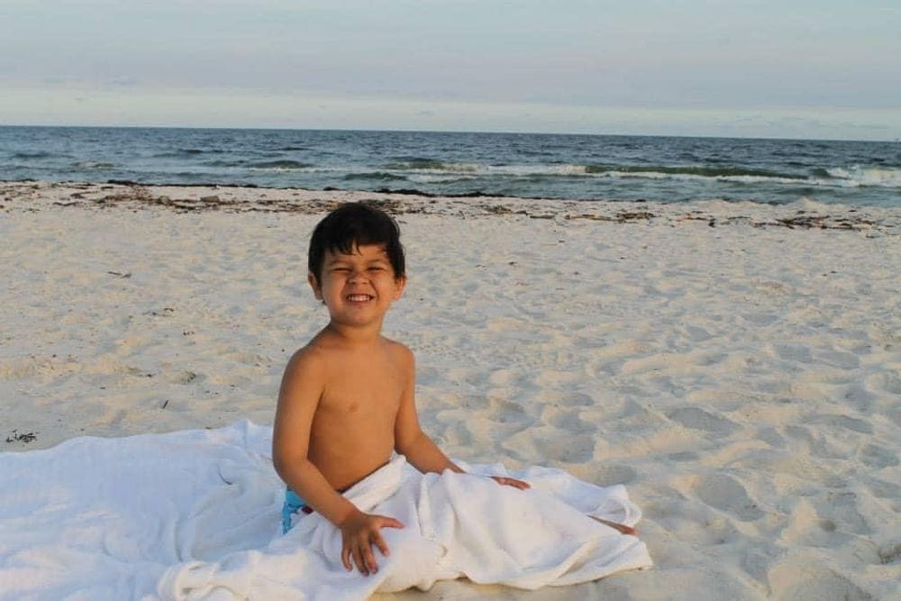 A young boy with a beach blanket around his lap smiles while enjoying a sunny day at Gulf Shores in Alabama, one of our recommended kid-friendly U.S. beaches for families.