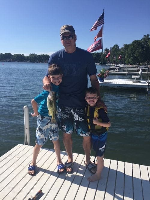 A dad and two kids stand on a dock over Lake Wawasee holding fresh caught fish.