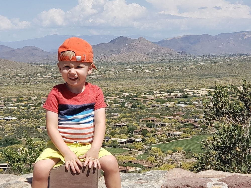 A young boy with a red cap on backwards sits on a stone fence with a desert view behind him while exploring Pinnacle Peak Park.