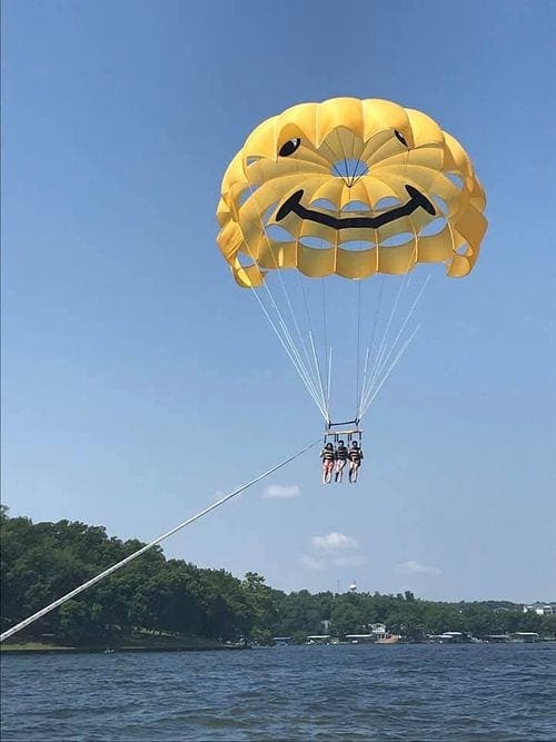 A yellow parasail holding three people flies behind a boat on Lake of the Ozarks.