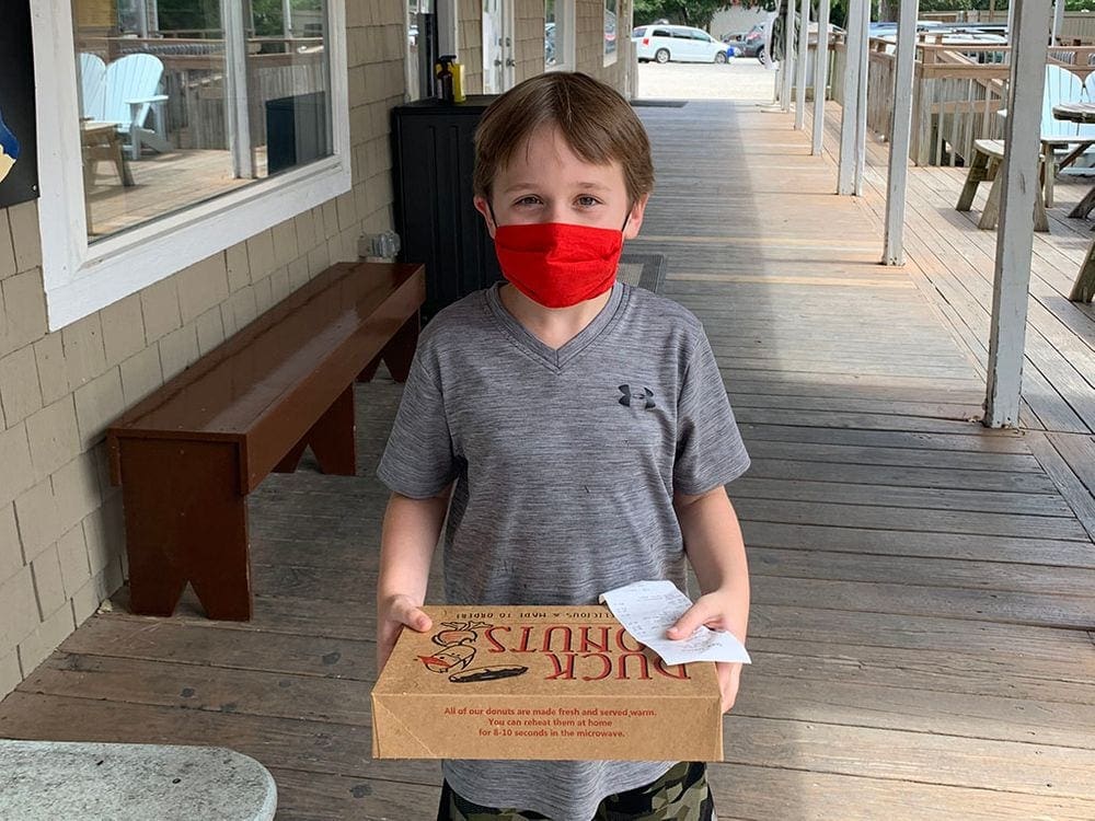 A young boy wearing a gray shirt and a red mask holds a box of Duck Donuts.