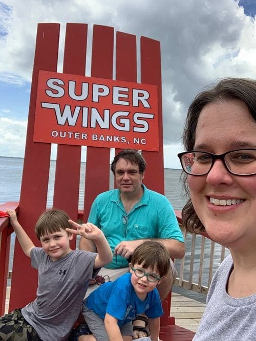 Parents and their two young boys take a selfie in front of a large red chair in the Outer Banks, a sign on the chair reads "Super Wings, Outer Banks, NC".
