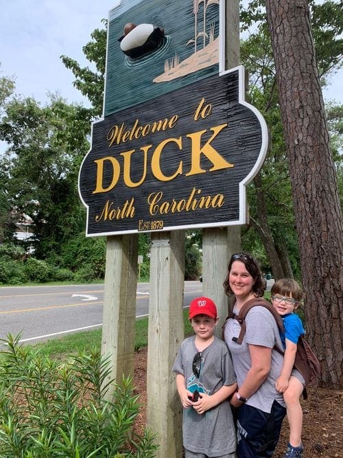 A mom, wearing one young son, and standing with the other, pose under a sign reading "Welcome to Duck, North Carolina".