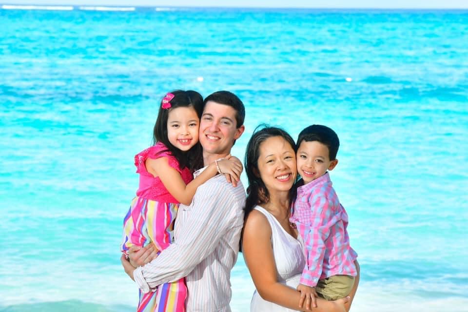A Asian family of four poses while standing with crystal blue waters behind them on a beach.