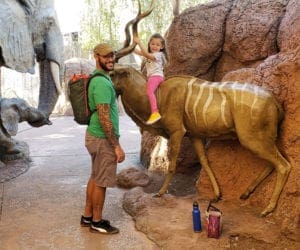 A dad stands near his young daughter as she rides a statue of an African animal at the Phoenix zoo.