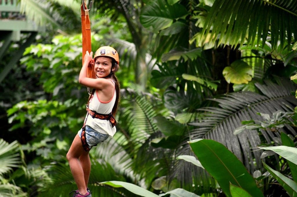 A young girl zip-lines among lush foliage in Hawaii.