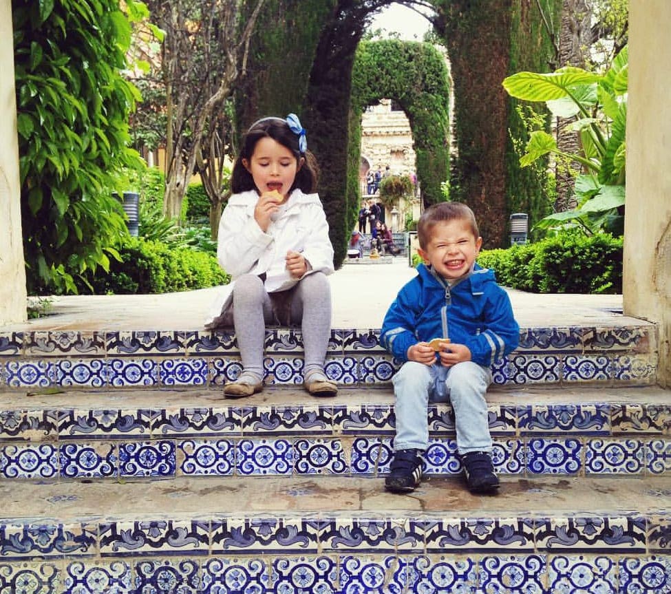Two kids sit smiling on the steps within Alcazar Palace.