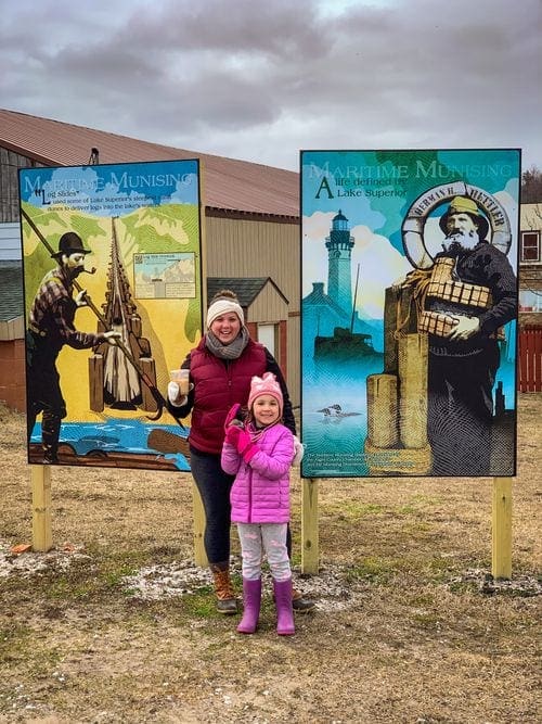 A mom and her daughter stand between two historical signs in Munising, one of the best places to visit in Michigan’s Upper Peninsula with kids.