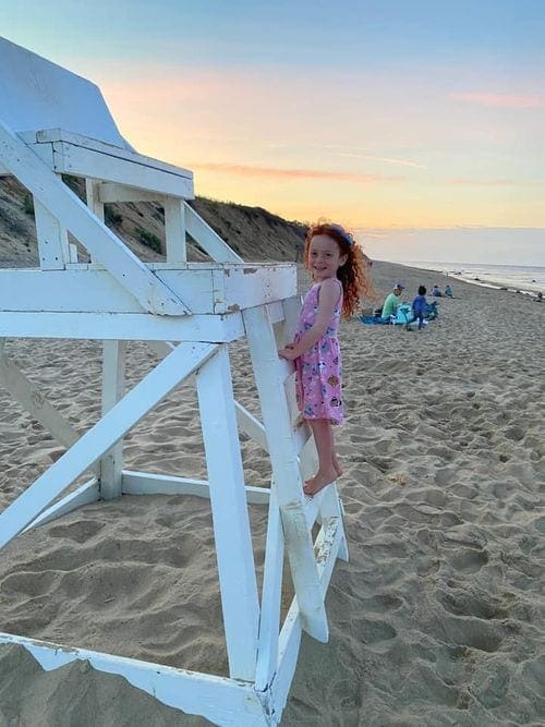 A young girl with red hair and a pink dress climbs a life guard chair on Coast Gaurd Beach in Cape Cod.