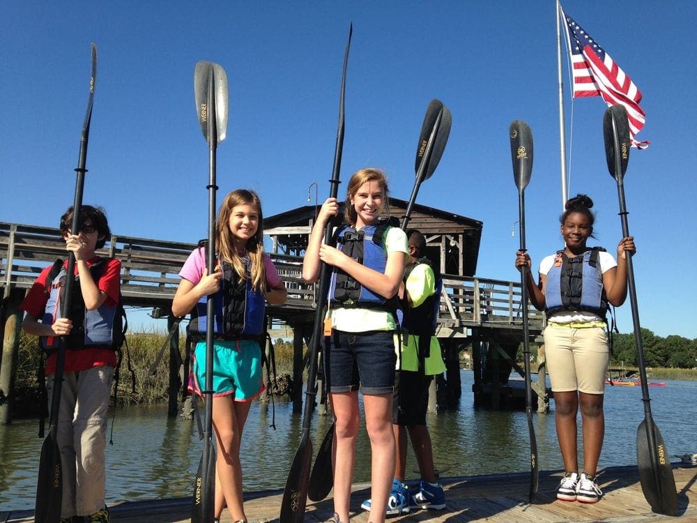 Six kids hold kayak paddles on a dock before a Coastal Expeditions tour.
