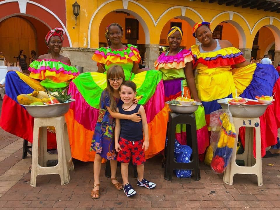 Two small American children stand in the center of four Colombian women in colorful dresses with baskets of spice and produce.